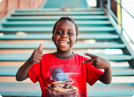 Smiling child on outdoor steps giving two thumbs up, representing people helped by Worldcoo