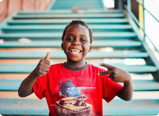 Smiling child on outdoor steps giving two thumbs up, representing people helped by Worldcoo