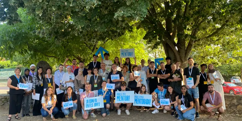 Foto grupal al aire libre de muchas personas sonriendo y mostrando carteles que dicen "Yo confío en los jóvenes" y "¡Viva mi mentora!"