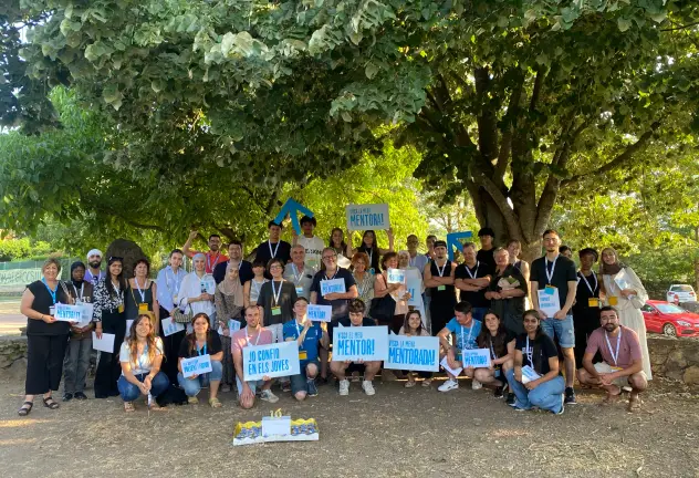 Foto grupal a l'aire lliure de moltes persones somrient i mostrant cartells que diuen "Jo confio en els joves" i "Visca la meva mentora!"