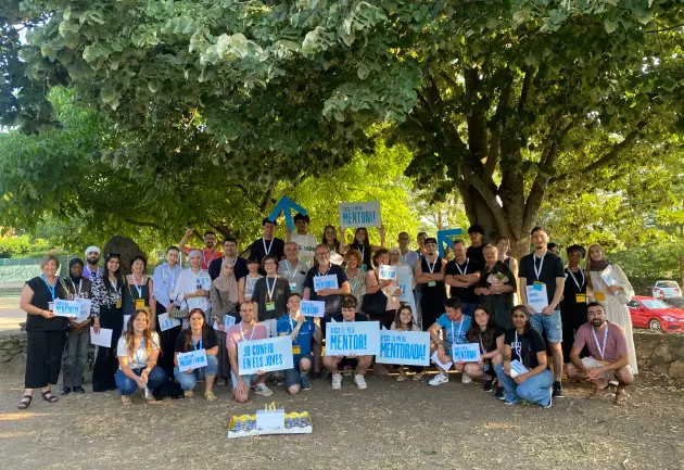 Foto grupal a l'aire lliure de moltes persones somrient i mostrant cartells que diuen "Jo confio en els joves" i "Visca la meva mentora!"